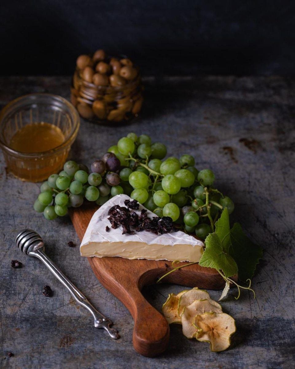 A wedge of Van Gaalen Camembert on a wooden board with grapes, honey and olives